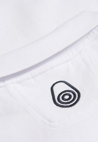 Nahaufnahme eines weißen Baumwollhemdes mit einem schwarzen gestickten ovalen Logo mit kreisförmigen Linien und einer strukturierten Rippkragen.