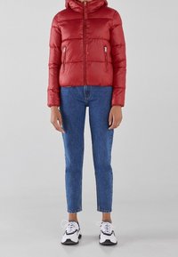 Rød pufferjakke med hætte, lynlås foran og sidelommer. Bæres med blå jeans og sort-hvide sneakers.