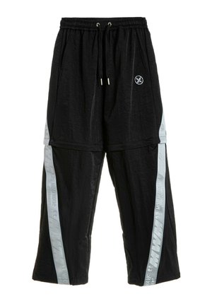 Joven con rastas que lleva una camiseta blanca extragrande y pantalones negros holgados con rayas grises en los lados, de pie frente a un fondo blanco liso.