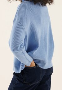 Person mit lockigem Haar, die einen locker sitzenden, hellblauen Pullover trägt, mit einer Hand in der Tasche einer dunklen Jeans, vor einem neutralen Hintergrund.