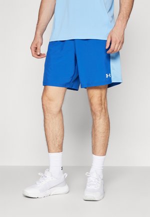 Homme portant un short de sport bleu, une chemise bleu clair, des chaussettes blanches et des chaussures de sport blanches, debout sur une surface claire.