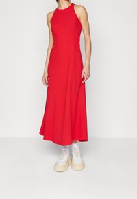 Robe maxi sans manches rouge avec un corsage ajusté, une jupe évasée et une texture lisse, associée à des baskets blanches à semelles épaisses.