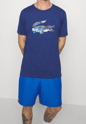 Mann trägt ein marineblaues T-Shirt mit einem mehrfarbigen Lacoste-Krokodil-Logo und leuchtend blauen Shorts, steht mit den Händen hinter dem Rücken.