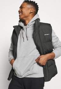 Vest puffer cu dungi verticale negre și gri, cu fermoar în față, textură matlasată, purtat peste un hanorac gri deschis cu buzunar tip kangaroo.