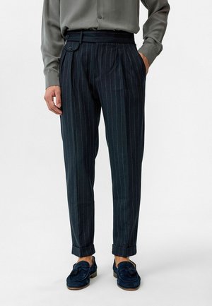 Donkerblauwe pinstrepenbroek met plooien en een haakje, gecombineerd met een grijs shirt. Heeft opgerolde manchetten en een slanke, op maat gemaakte pasvorm.