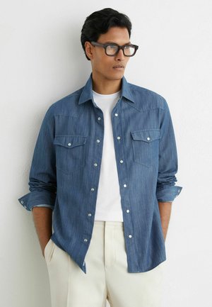 Mladý muž s čiernymi vlasmi a okuliarmi, oblečený v modrej rifľovej košeli cez biele tričko a krémové nohavice, stojaci pri bielej stene.