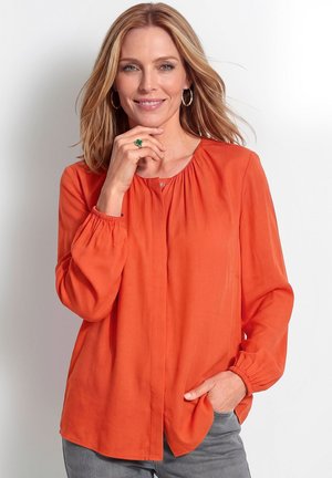 Frau mit schulterlangen blonden Haaren trägt eine orangefarbene Bluse mit langen Ärmeln, graue Hose, Creolen und einen grünen Kleeblatt-Ring, lächelt mit der Hand nahe am Gesicht.