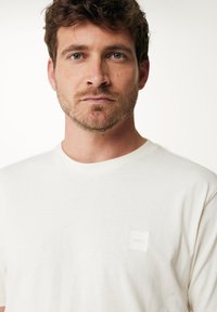 Bílý bavlněný tričko s krátkými rukávy a kulatým výstřihem, se malou čtvercovou nášivkou na levé straně hrudi. Hladká textura.