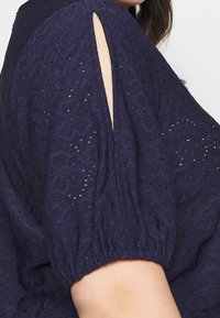 Tmavě modrá blůzka z texturované látky s květinovým vzorem vyřezávaným do látky, s krátkými nabíranými rukávy, bočním rozparkem a elastickými manžetami.