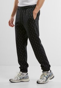 Svarte joggebukser med et diskret logomønster over hele, elastisk midje og ribbestrikket ankelkant, kombinert med blanke sølvfargede joggesko.