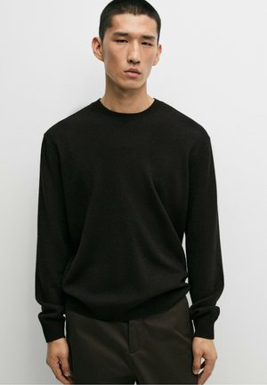 Uomo che indossa un maglione nero semplice a maniche lunghe e pantaloni scuri, in piedi davanti a uno sfondo chiaro uniforme, guarda avanti.