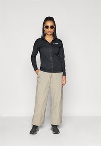 Μαύρο μπουφάν με φερμουάρ, μπεζ παντελόνι χαλαρής εφαρμογής και σκούρα υποδήματα. Μοντέλο που φοράει ανοιχτόχρωμα γυαλιά ηλίου. Λείο ύφασμα με απλό σχέδιο.