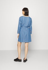 Vestido de ganga com mangas longas, cintura ajustada e gola redonda. Tecido azul com uma textura suave. Usado com tamancos pretos.