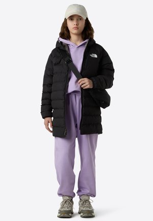 Giacca a vento nera con cappuccio, felpa viola e pantaloni della tuta coordinati. Cappello beige e sneakers grigie con suola texturizzata. Borsa a tracolla.