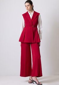 Punane kohandatud vest peplum-kujuga, koos laiade punaste pükstega. Valge kraeäärtega särk all, millel on vöödiga detail ja sile kangas.