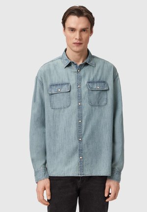 Ung mand iført en lyseblå denimskjorte med knapper og to forlommer samt sorte bukser, stående foran en ensfarvet baggrund.