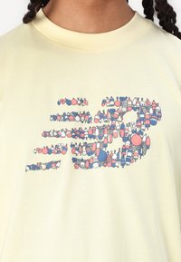Světle žluté bavlněné tričko s vícebarevným grafickým vzorem, připomínající různé tvary a postavy, umístěné na přední části.