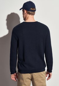 Pull en maille bleu marine avec un motif texturé, col rond et manches longues. Porté avec un pantalon beige, vu de dos.