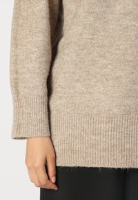 Beige kötött pulóver, amely lágy tapintású, bordás mandzsettákkal és aljjal rendelkezik. Az anyag finom mintázatú, jól látható szálakkal az extra részletességért.