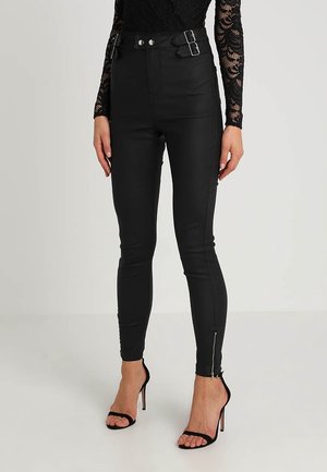 Femme portant un pantalon noir taille haute avec fermetures éclair latérales et détails de boucle, associé à un top en dentelle noire et des sandales noires à talons hauts.