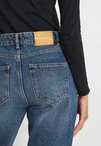 Hög midja blå denimjeans med en beige läderetikett, med två bakfickor och kontrasterande sömmar, visade från sidan.