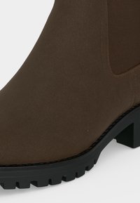 Bottine en daim marron foncé avec semelle noire épaisse et empiècement latéral élastique, montrée en gros plan.