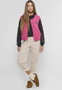 Roze fleece vest met knopen over een zwarte langemouwen shirt, gecombineerd met lichtbeige broek en bruine slippers, met crèmekleurige sokken.