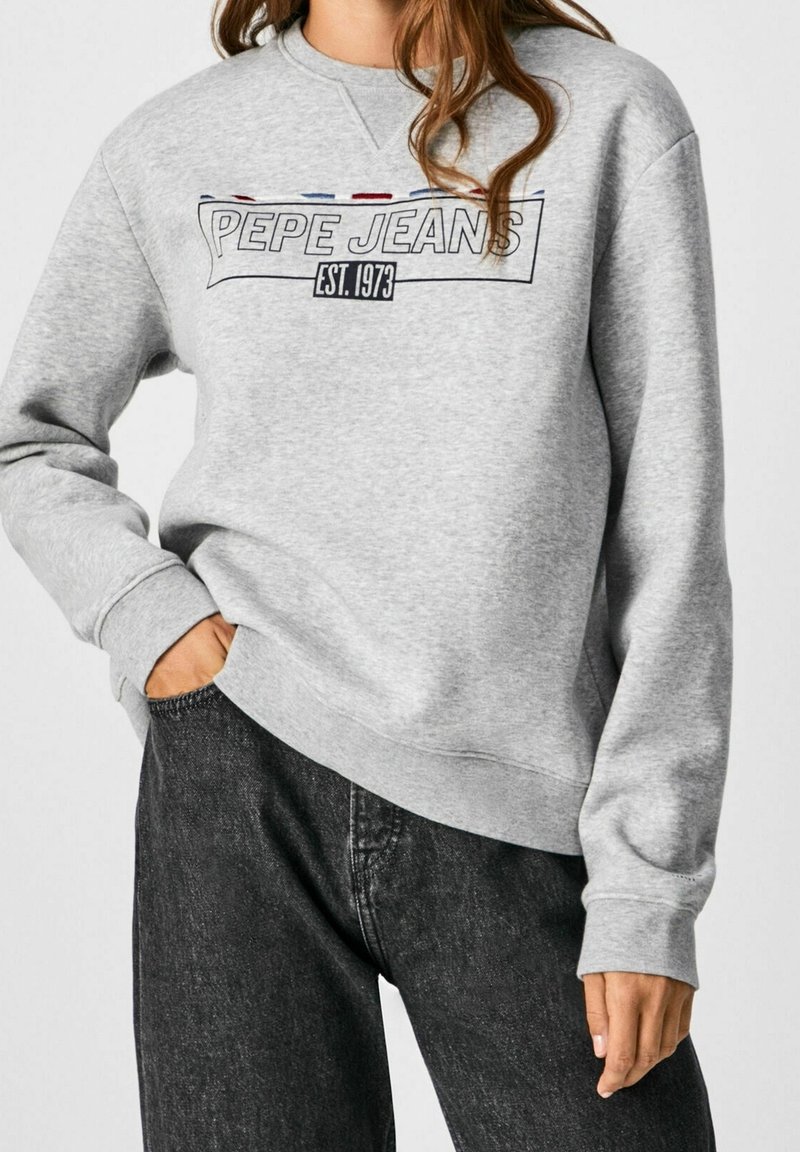 Personne portant un sweat-shirt gris clair avec le texte "PEPE JEANS EST. 1973" et un jean gris foncé, main dans la poche sur la hanche.