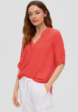 Koraalkleurige blouse met een V-hals, korte opgerolde mouwen en subtiele plooien. Gecombineerd met witte, trekkoordbroeken van lichtgewicht stof.