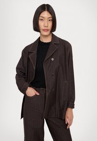 Veste marron à rayures, avec un col cranté, deux poches avant et des coutures orange contrastantes ; associée à un haut noir à côtes.