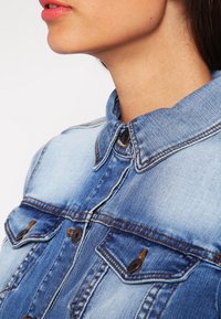 Džínová bunda ve světle modré barvě, s knoflíkovým zapínáním vpředu, límcem košilového typu, dvěma náprsními kapsami a viditelnými švy.