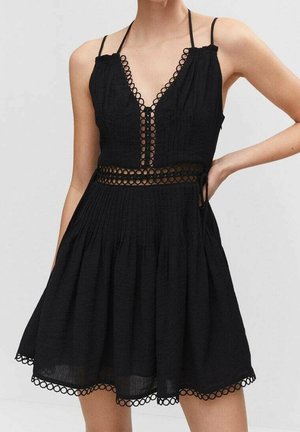 Kobieta w czarnej sukience bez rękawów z ozdobą z okrągłej koronki na dekolcie, w talii i u dołu sukienki, stoi z jedną ręką na biodrze.