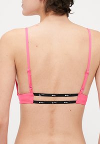 Roze sportbeha met dunne banden, voorzien van een zwarte band met witte Nike-logo's op de achterkant. Smooth textuur, minimalistisch ontwerp, verstelbare banden.