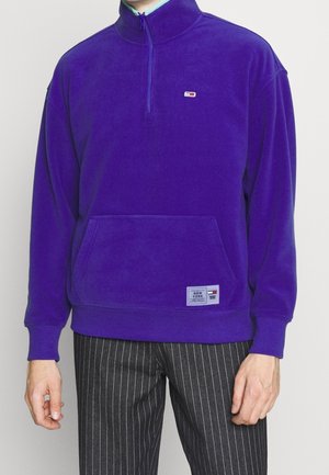 Mand iført lilla kvart-lynlåsfleece med frontlomme og lille logo, kombineret med sorte bukser med nålestriber.