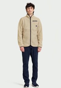 Bež flis jakna s patentnim zatvaračem sprijeda, s džepom na prsima i tamnim detaljima. U kombinaciji s mornarsko plavim hlačama i sivoj tenisicama.
