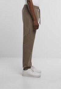 Beige Jogginghose mit elastischem Bund und Kordelzug, versehen mit einem vertikalen Logo-Detail an der Seite, kombiniert mit weißen Turnschuhen.
