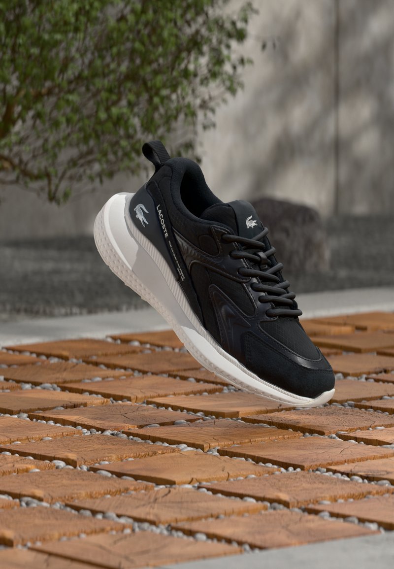 Baskets Lacoste noires avec semelle blanche flottant au-dessus de carreaux en bois en extérieur, présentant le logo Lacoste et une fermeture à lacets.