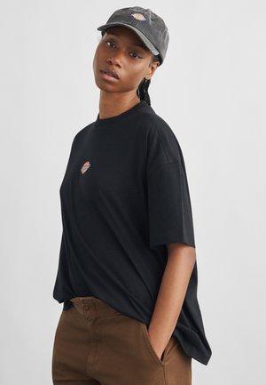 Fiatal személy fekete logós pólóban és szürke sapkában, barna nadrágban, egyik kezét a zsebében tartva, egyszínű háttér előtt áll.