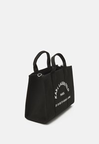 Musta kangaskassi tukevilla kahvoilla ja kohokuvioidulla valkoisella tekstillä, jossa lukee "KARL LAGERFELD PARIS" ja osoitetiedot. Suorakulmainen muoto.