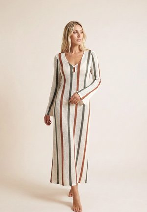 Mujer que lleva un vestido maxi de manga larga y cuello en V con rayas verticales en óxido, gris y blanco, de pie descalza sobre un fondo neutro.