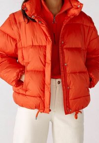 Veste matelassée rouge avec un col montant, des boutons-pression et des cordons de serrage réglables à l'ourlet. Texture matelassée ; associée à un haut en maille rouge.