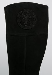 Bota de ante negra hasta la rodilla con un logotipo en relieve cerca de la parte superior, con una textura suave y un diseño minimalista.