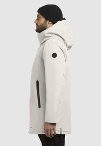 Lys grå, polstret parka med stor hætte, sort lynlås lomme og logo patch på ærmet. Fremstillet af vejrbestandigt materiale.