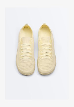 Pár lehkých světlounce žlutých pletených tenisek s texturovanými podrážkami a sladěnými tkaničkami, zobrazený shora na bílém pozadí.