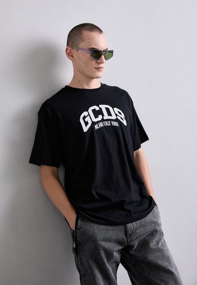 T-shirt nera in cotone con stampa bianca "GCDS MILANO ITALY 1988", abbinata a pantaloni in denim grigio. Il modello indossa occhiali da sole angolari con tonalità verde.