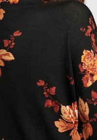 Czarny sweter z dużymi pomarańczowymi i kremowymi wzorami kwiatowymi oraz zielonymi akcentami. Materiał wydaje się być dziany i ma miękką teksturę.
