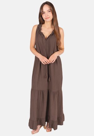 Frau mit langen braunen Haaren, die ein ärmelloses braunes Maxikleid mit Rüschen und Quastenbändern trägt, steht und lächelt vor einem schlichten Hintergrund.