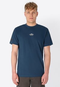 Junger Mann mit lockigem Haar, der ein navyblaues T-Shirt mit einem kleinen weißen Flugzeuglogo und Text in der Mitte der Brust trägt.