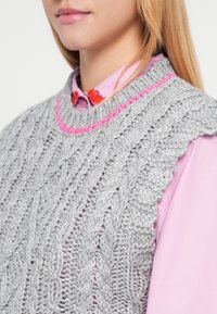 Szary dziergany kamizel z warkoczowym wzorem, z okrągłym dekoltem podkreślonym różowym obszyciem, noszony na różowej koszuli z kołnierzykiem.