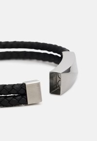 Pulsera de cuero trenzado negro con un cierre de metal plateado liso que tiene un extremo cuadrado y una sección curva, destacando las texturas contrastantes.